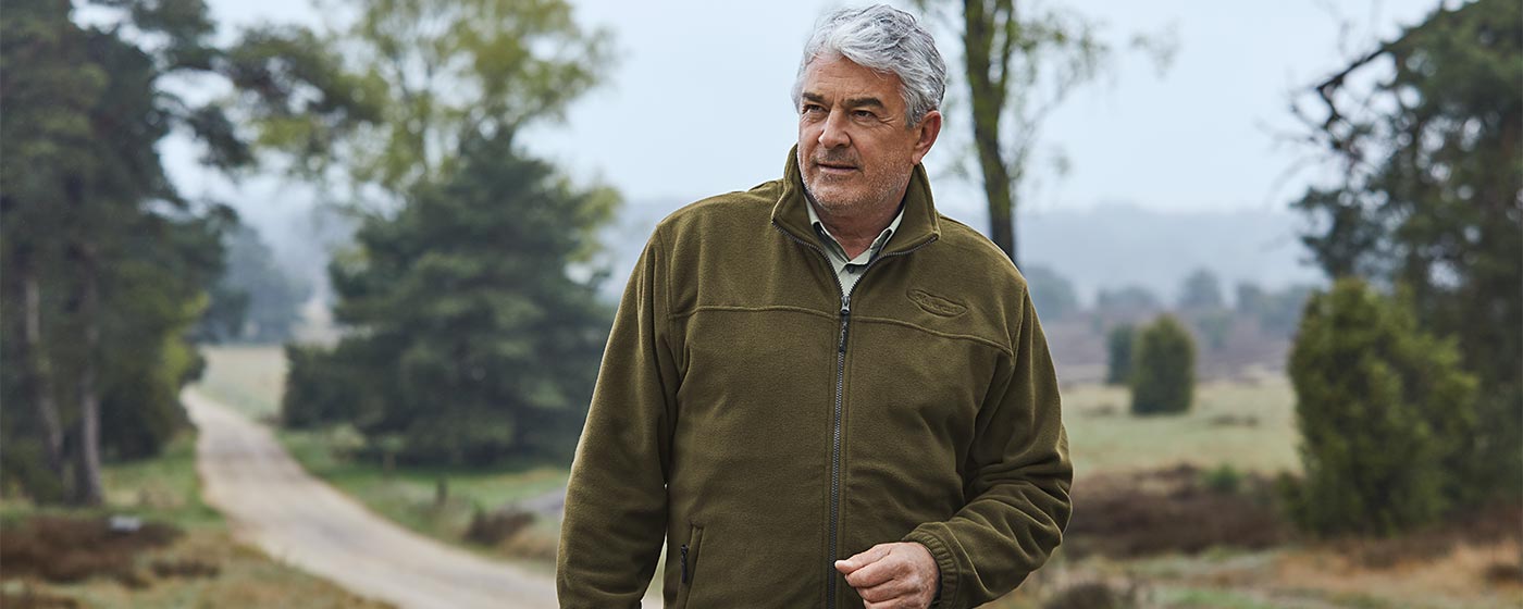 „Älterer Mann in olivgrüner Outdoorjacke geht einen Naturweg entlang und blickt gedankenverloren zur Seite; im Hintergrund liegen Bäume und offene Heideflächen.