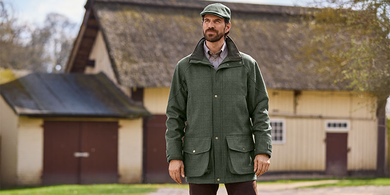 Mann in grüner Outdoorjacke und passender Schirmmütze steht vor einem alten Gebäude mit Reetdach und Scheunentoren und blickt leicht zur Seite.